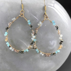 May include: A pair of gold-toned hoop earrings. Each earring is adorned with small, colorful beads in shades of blue, black, white, and pink. The earrings have a teardrop shape and are suspended from gold-colored ear wires.