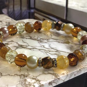 May include: A beaded bracelet featuring a variety of amber, gold, and pearl-colored beads. The bracelet includes faceted and round beads, with silver-toned spacers. The bracelet is displayed on a marble-patterned surface.
