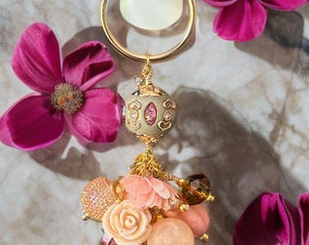 Pode incluir: Brinco dourado com um pendente decorativo com uma borla de contas rosa e dourada, flores rosa e uma conta redonda rosa. O brinco é exibido com flores rosa brilhantes em um fundo de mármore.