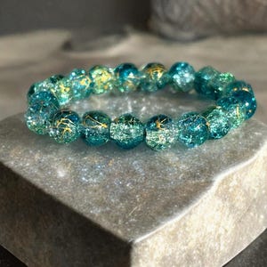 Pode incluir: Uma pulseira de contas turquesa e douradas. A pulseira apresenta contas redondas com efeito craquelado em tons de turquesa e dourado. As contas são enfiadas para formar um círculo completo. A pulseira é exibida em uma superfície de pedra cinza.