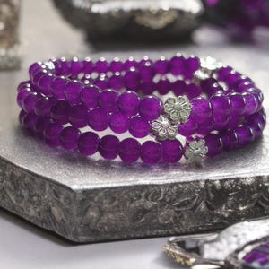 May include: A multi-strand bracelet composed of deep purple, translucent, spherical beads. The bracelet features three distinct silver-toned metal flower-shaped spacers interspersed along the circular strand, creating a layered appearance.