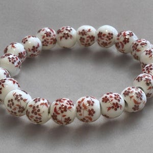 Pode incluir: Uma pulseira de contas brancas com um padr&atilde;o repetido de desenhos florais vermelhos. A pulseira &eacute; feita de contas redondas, cada uma adornada com um delicado motivo floral. As contas s&atilde;o enfiadas para formar um c&iacute;rculo completo.
