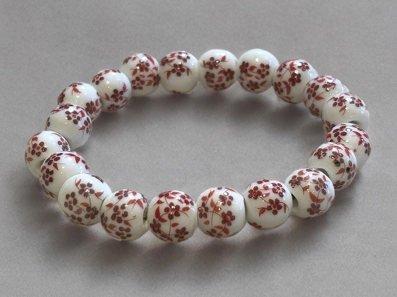 Pode incluir: Uma pulseira de contas brancas com um padr&atilde;o repetido de desenhos florais vermelhos. A pulseira &eacute; feita de contas redondas, cada uma adornada com um delicado motivo floral. As contas s&atilde;o enfiadas para formar um c&iacute;rculo completo.