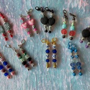 May include: A collection of handmade earrings with various designs. The earrings feature beads in colors like red, pink, blue, black, and green. Some have heart-shaped beads, while others have decorative metal accents. The earrings are displayed on a light blue surface.