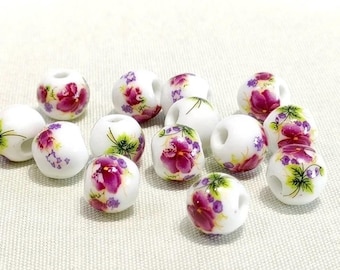 May include: White ceramic beads with a floral design featuring purple and pink flowers and green leaves. The beads are round with a hole through the center, suitable for jewelry making or crafts. The beads are scattered on a white surface.