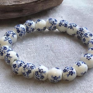 Pode incluir: Uma pulseira de cer&acirc;mica branca com padr&otilde;es florais azuis. As contas redondas s&atilde;o enfiadas, criando um design circular. A pulseira &eacute; exibida em uma superf&iacute;cie de cor clara.