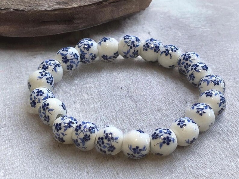 Pode incluir: Uma pulseira de cer&acirc;mica branca com padr&otilde;es florais azuis. As contas redondas s&atilde;o enfiadas, criando um design circular. A pulseira &eacute; exibida em uma superf&iacute;cie de cor clara.