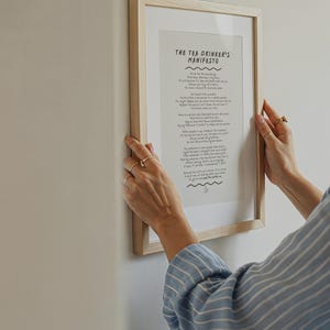 Peut inclure: Une affiche encadrée intitulée "The Tea Drinker's Manifesto" avec du texte noir sur fond blanc. Le cadre est en bois clair. L'affiche est tenue contre un mur blanc.