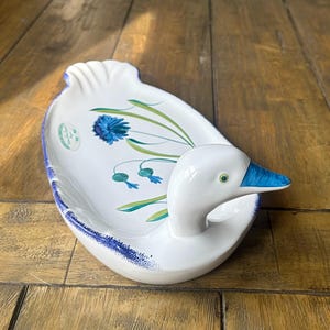 May include: White ceramic duck-shaped serving dish with a blue beak and green eyes. The dish features a floral design with blue flowers and green leaves. The rim is trimmed with blue paint.