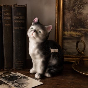 May include: A ceramic cat figurine with a glossy finish, featuring a white chest and face with grey shading on the back and ears. The cat is in a seated position, looking upward. A small white label on the side of the cat displays the text Kat 2021.