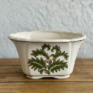 May include: Cream-colored ceramic planter with a scalloped rim and a flared, octagonal shape. The planter features a green and red floral design on the front. The rim is outlined with a thin brown line. The planter sits on a wooden surface.