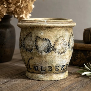 May include: A beige ceramic pot with a weathered, distressed finish featuring a band of embossed leaf patterns around the upper section and the word MULBERRY stamped in dark, recessed lettering along the lower base of the rounded vessel.