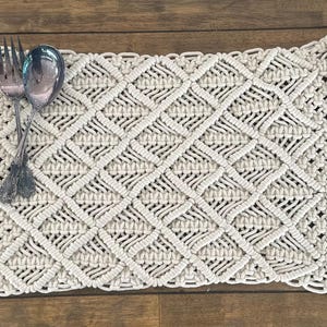 May include: Cream-colored macrame placemat with a diamond pattern and fringe edges. A fork and spoon rest on the placemat, which is displayed on a wooden table. The placemat is approximately 12 inches wide and 18 inches long.