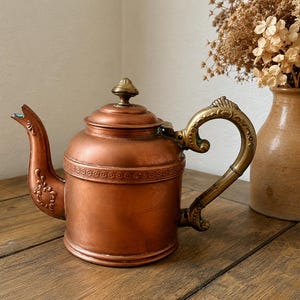 May include: A copper teapot featuring a brass handle and a matching brass lid finial. The body of the teapot is adorned with a decorative embossed floral band around the upper section, while the curved spout includes intricate relief detailing.