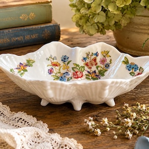 May include: White ceramic serving dish with a scalloped edge and floral design. The dish features a raised, footed base and is decorated with colorful flowers in shades of red, blue, yellow, and purple. The dish is on a wooden surface.