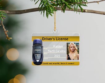 May include: A clear plastic ornament shaped like a driver's license. The ornament features a photo, a car graphic, and the text "Driver's License." The text includes the name "Raiden," "North Carolina," and "2025." The bottom text reads "Look out world, here I come!"