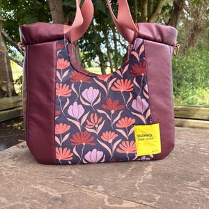 Op de afbeelding: Een draagtas met een donkerpaars bloemenpatroon van roze en lichtpaarse bloemen, geaccentueerd door effen donkerpaarse zijpanelen en roze stoffen hengsels. Een felgeel vierkant label op de voorzijde bevat de tekst EIGENWIJS. met trots.