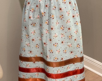 May include: A light blue skirt with an elastic waistband and a floral pattern. The skirt features horizontal ribbons in shades of brown, red, and orange near the hem. The fabric is decorated with small flowers and white dots.