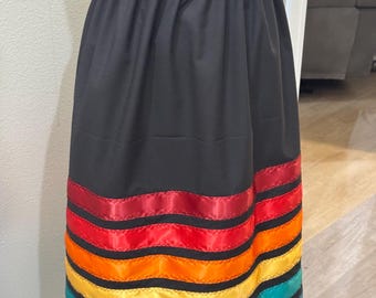 May include: A black skirt with a ruffled waistband and a series of horizontal, colorful ribbons. The ribbons are in shades of red, orange, yellow, teal, and blue, creating a vibrant rainbow effect. The skirt appears to be made of a smooth fabric.