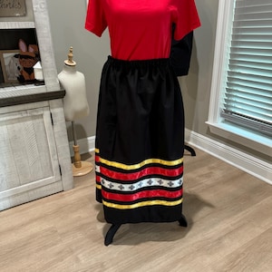 May include: A red short-sleeved top paired with a black skirt. The skirt features horizontal bands of yellow, red, and white ribbon. The outfit is displayed on a mannequin, showcasing a traditional or cultural garment.