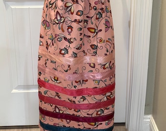 May include: A pink skirt with a floral pattern and colorful ribbon accents. The skirt features a repeating pattern of stylized flowers and leaves in shades of blue, yellow, and orange. Horizontal ribbons in pink, red, and teal add a decorative touch to the skirt.