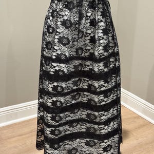 May include: A long black lace skirt with a floral pattern. The skirt has a scalloped hemline and horizontal bands of lace. The skirt is shown on a mannequin.