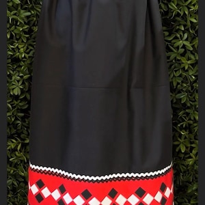 May include: A black skirt with an elasticized waistband and a red horizontal band near the hem. The red band features a repeating pattern of white and black diamonds, bordered by white zigzag trim along the top and bottom edges.