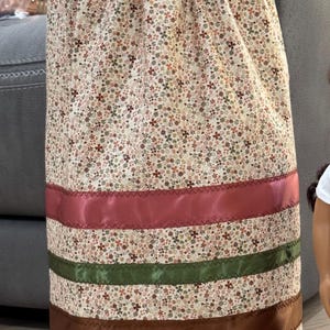 May include: A floral print skirt with a beige background and small red, green, and brown flowers. The skirt has three horizontal satin ribbons in red, green, and brown. The skirt is displayed on a wooden stand.