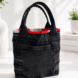 May include: Black denim tote bag with two black handles. The bag has a red and white striped lining and a pocket on the front. The bag is made of recycled denim.