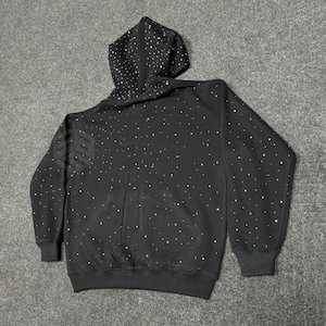 Op de afbeelding: Zwarte hoodie met een wit sterrenpatroon. De sweatshirt heeft een capuchon met trekkoord, lange mouwen en geribde manchetten en zoom. Het ontwerp heeft een verspreid patroon van kleine witte stippen over de stof.