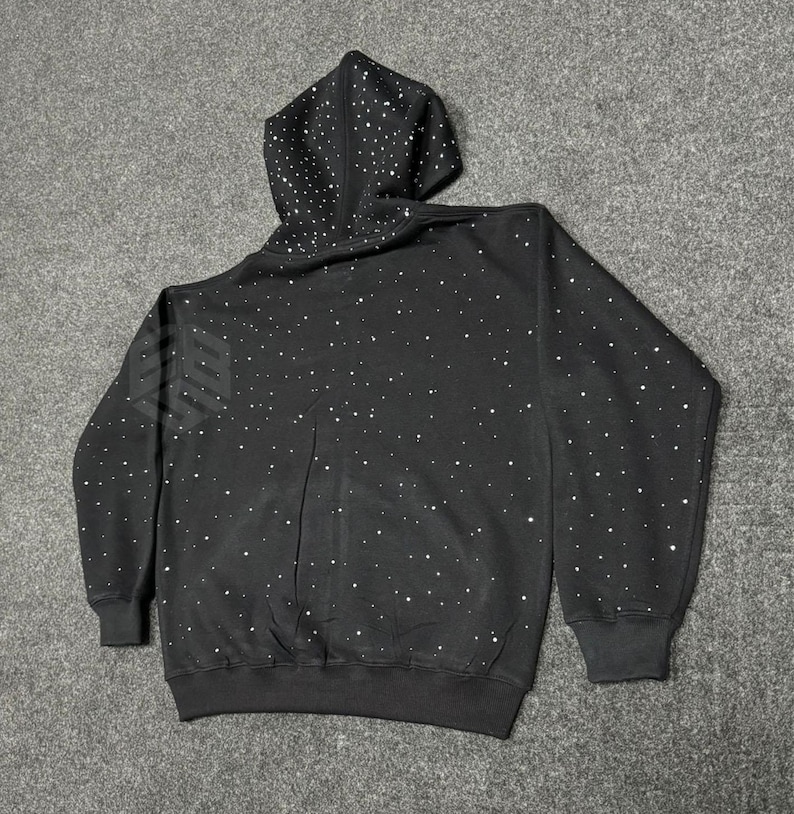 Op de afbeelding: Zwarte hoodie met een wit sterrenpatroon. De sweatshirt heeft een capuchon met trekkoord, lange mouwen en geribde manchetten en zoom. Het ontwerp heeft een verspreid patroon van kleine witte stippen over de stof.
