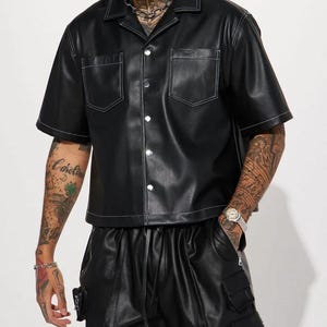 May include: Black leather shirt with white stitching detail, short sleeves, and two front pockets. The shirt has a button-up closure and is paired with black leather shorts. The outfit is a modern take on a classic style.
