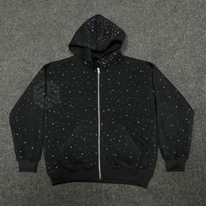 Op de afbeelding: Zwarte hoodie met ritssluiting en een sterrenbeeldpatroon van witte stippen. De hoodie heeft een capuchon met trekkoord, een volledige ritssluiting en twee voorzakken. De manchetten en zoom zijn geribbeld.