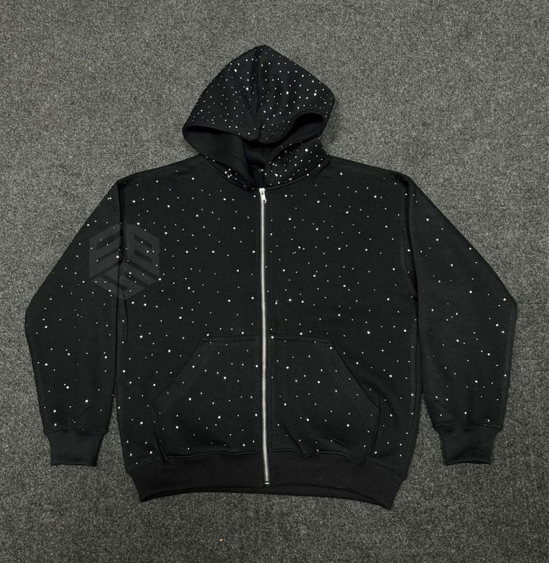 Op de afbeelding: Zwarte hoodie met ritssluiting en een sterrenbeeldpatroon van witte stippen. De hoodie heeft een capuchon met trekkoord, een volledige ritssluiting en twee voorzakken. De manchetten en zoom zijn geribbeld.