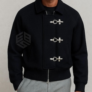 Puede incluir: Chaqueta negra con cuello y tres cierres de palanca plateados en la parte delantera. La chaqueta tiene cierre de cremallera y puños de canalé. La chaqueta está hecha de un material grueso.