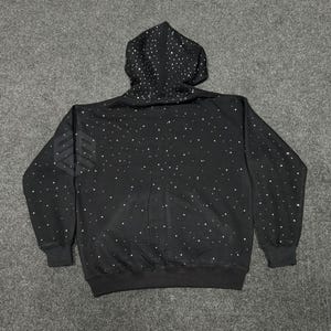 Op de afbeelding: Zwarte hoodie met een patroon van witte stippen, die op sterren lijken. De hoodie heeft een capuchon met trekkoord, lange mouwen en een geribbelde zoom en manchetten. Een wit logo is zichtbaar op de linkermouw.