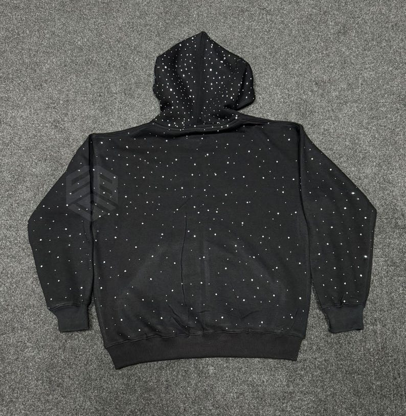 Op de afbeelding: Zwarte hoodie met een patroon van witte stippen, die op sterren lijken. De hoodie heeft een capuchon met trekkoord, lange mouwen en een geribbelde zoom en manchetten. Een wit logo is zichtbaar op de linkermouw.