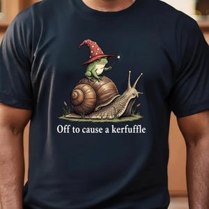 Könnte beinhalten: Dunkelblaues T-Shirt mit einer Fantasy-Illustration einer Schnecke und eines Frosches, die beide Zaubererhüte tragen. Der Text "Off to cause a kerfuffle" ist darunter gedruckt. Ein lustiges, skurriles Design.
