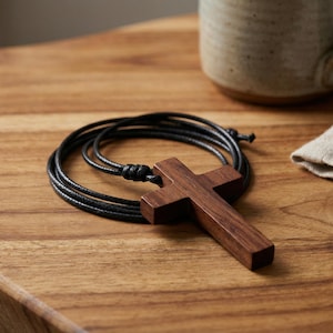 May include: A wooden cross pendant on a black cord necklace. The cross is a dark brown color and is lying on a wooden surface. A beige mug and a linen cloth are in the background.