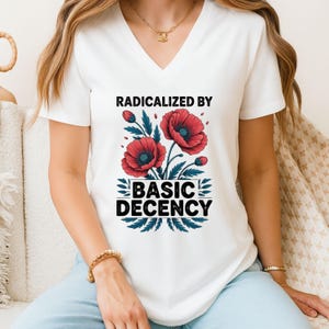 May include: White V-neck t-shirt with the text "RADICALIZED BY BASIC DECENCY" in black. The design includes red poppy flowers with green leaves. The shirt is a casual wear item.