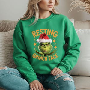 May include: Green sweatshirt featuring a graphic of the Grinch wearing a Santa hat, with the text "RESTING GRINCH FACE" in orange. The design includes yellow stars. The sweatshirt is a crew neck style.