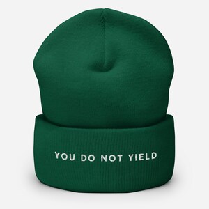 May include: A dark green beanie with the embroidered text "YOU DO NOT YIELD" in white. The hat is a folded-cuff style, made of a soft, knitted material. The beanie is a solid color, with a simple, classic design.