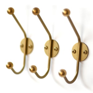 May include: Three gold metal hooks with rounded ends mounted on a white wall. Each hook has a circular base with two visible screws.