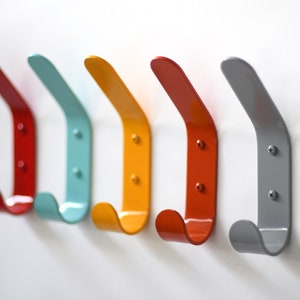 May include: Five metal hooks in different colors: red, turquoise, yellow, orange, and gray. The hooks are mounted on a white wall and are shaped like a curved letter J.