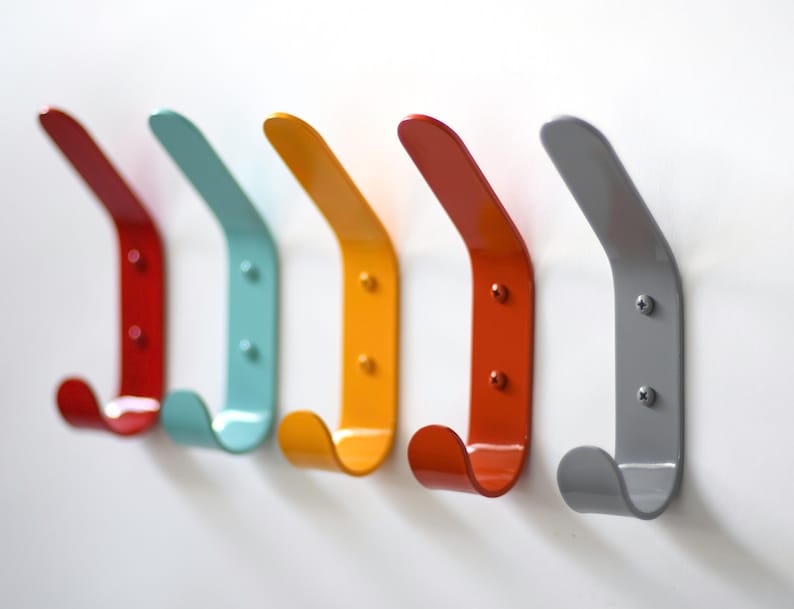 May include: Five metal hooks in different colors: red, turquoise, yellow, orange, and gray. The hooks are mounted on a white wall and are shaped like a curved letter J.