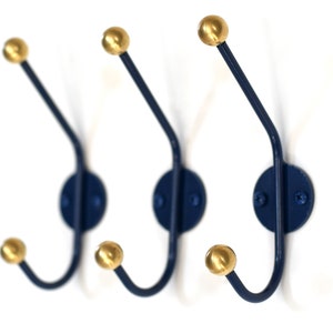 May include: Three navy blue metal hooks with gold ball finials. The hooks are mounted on a white wall.