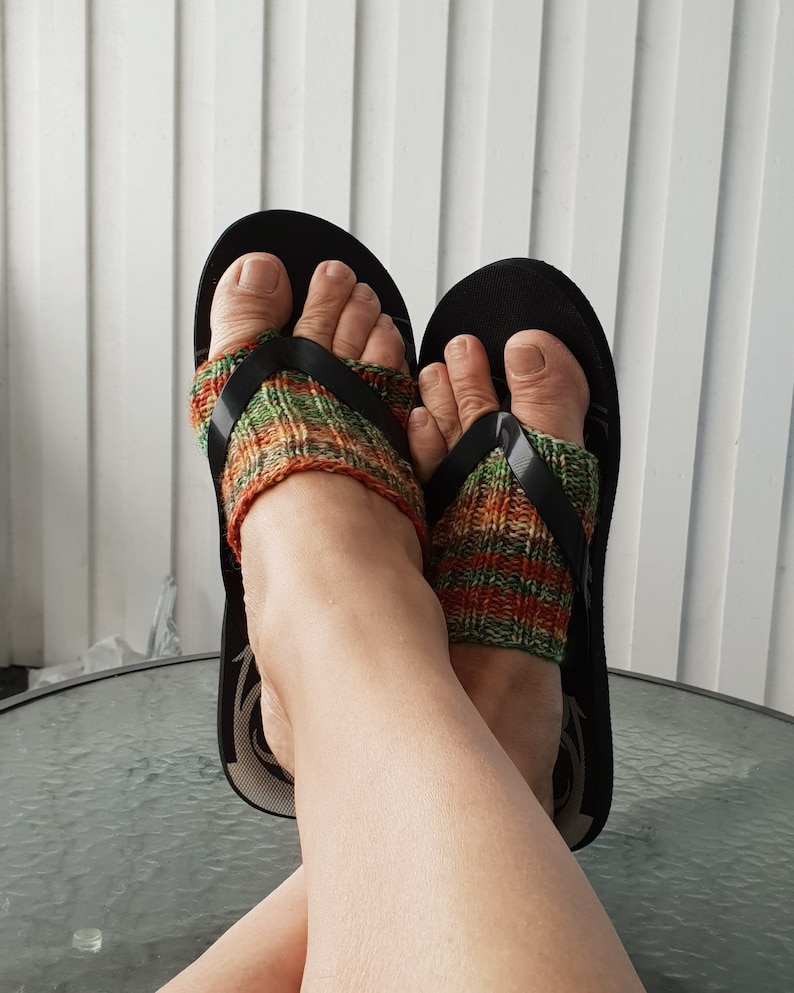 K&ouml;nnte beinhalten: Ein Paar schwarze Flip-Flops mit gestrickten Socken in Gr&uuml;n, Orange und Braun. Die Socken werden an den F&uuml;&szlig;en getragen und die Flip-Flops sind an den F&uuml;&szlig;en.