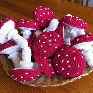 Puede incluir: Una cesta llena de setas de ganchillo rojas y blancas. Las setas tienen tallos blancos y sombreros rojos con puntos blancos.