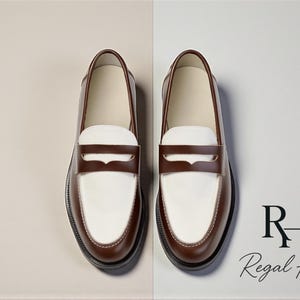 May include: A pair of two-tone leather loafers. The shoes have a white vamp and a brown saddle strap and trim. The soles are black. The brand name "Regal Hide" is displayed in the lower right corner.