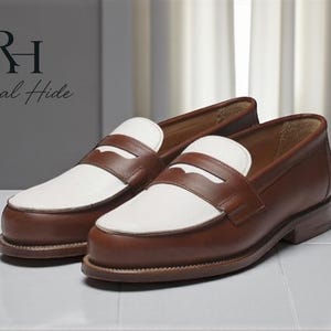 Op de afbeelding: Een paar bruin en witte leren penny loafers. De schoenen hebben een klassiek ontwerp met een bruin leren bovenwerk, witte wreef en een penny band. De schoenen worden op een wit oppervlak gepresenteerd.